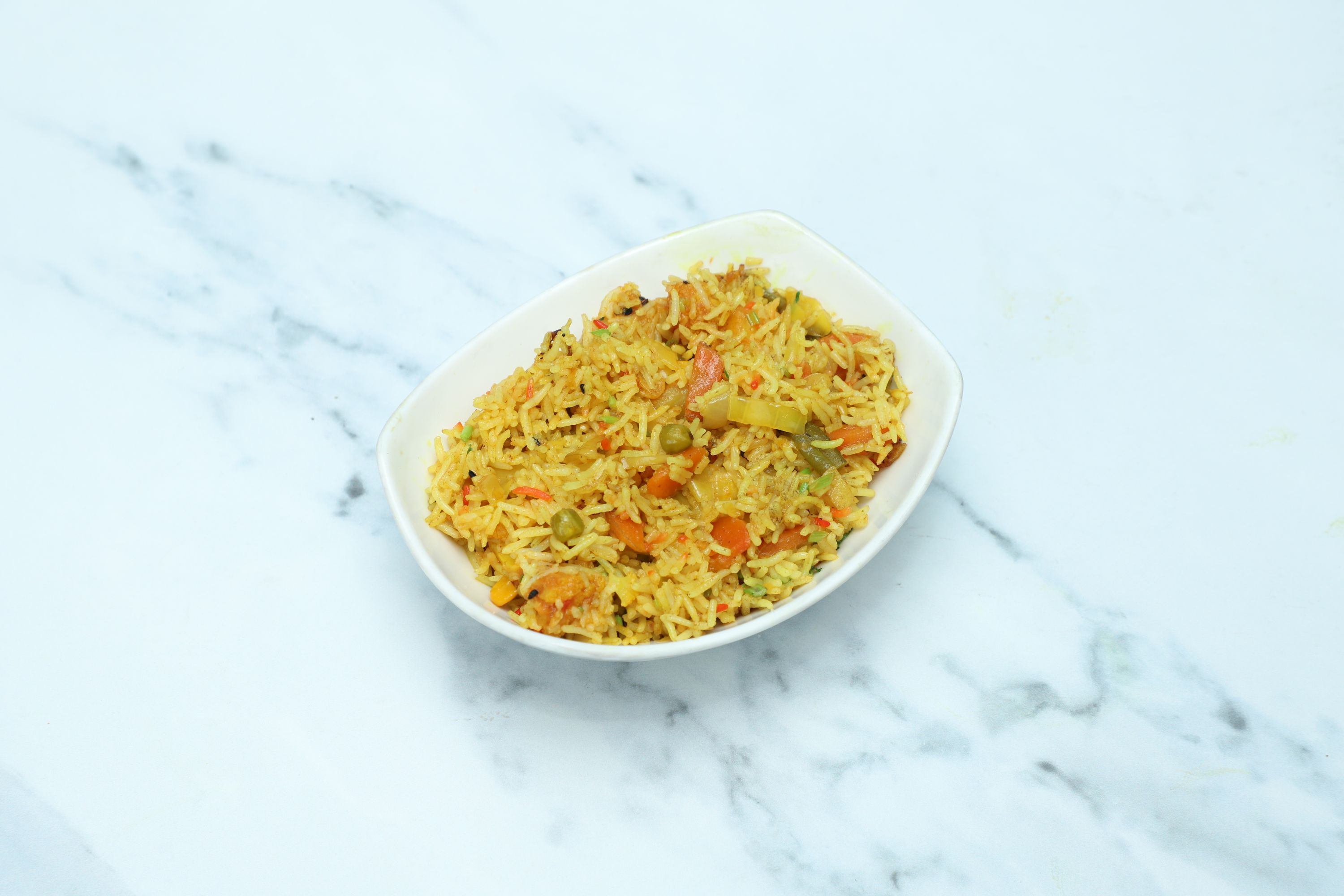 A white oval bowl filled with colourful vegetable biryani rice on a white marble surface