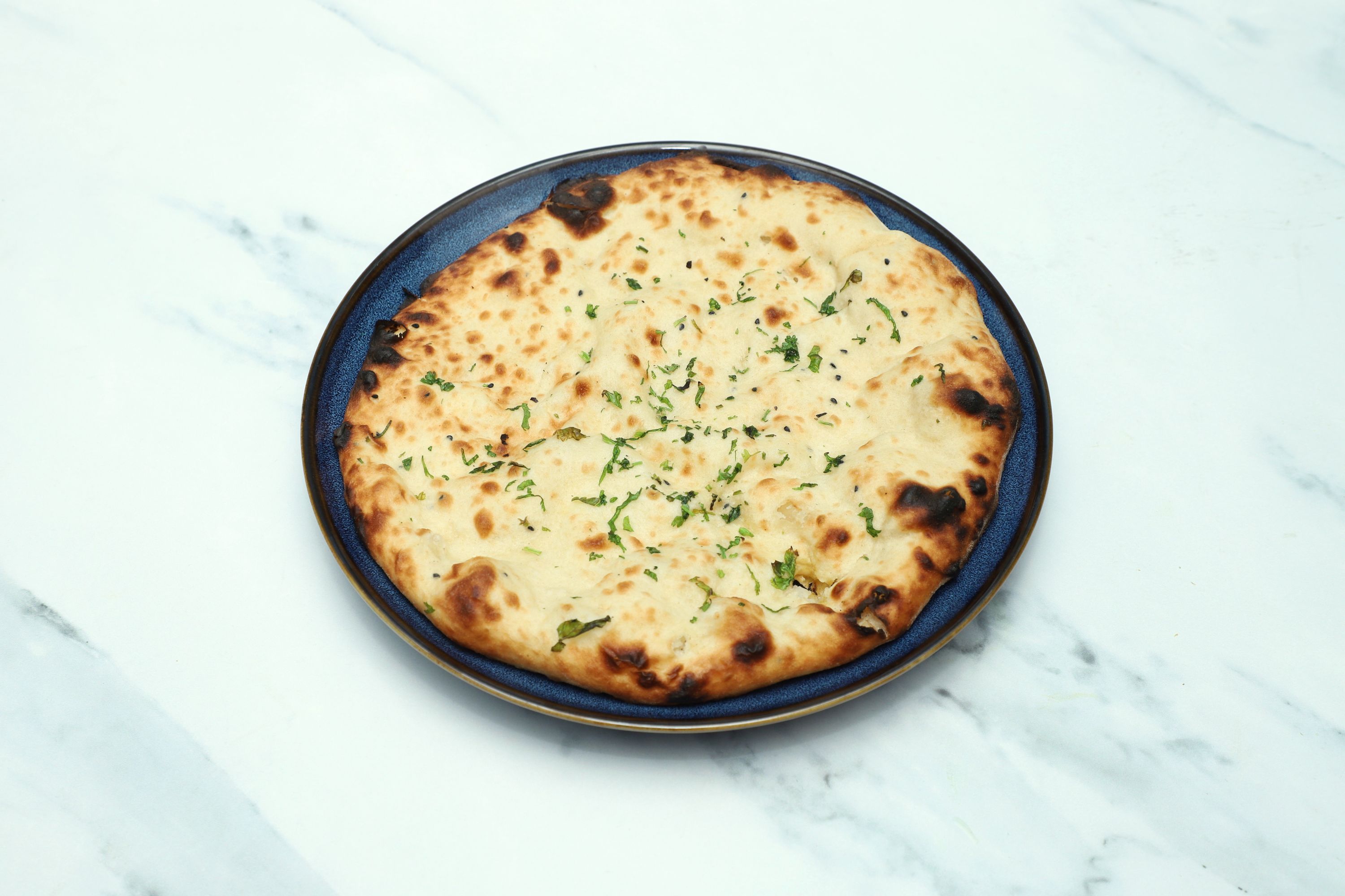 A round naan bread topped with fresh green herbs and sesame seeds on a blue plate.