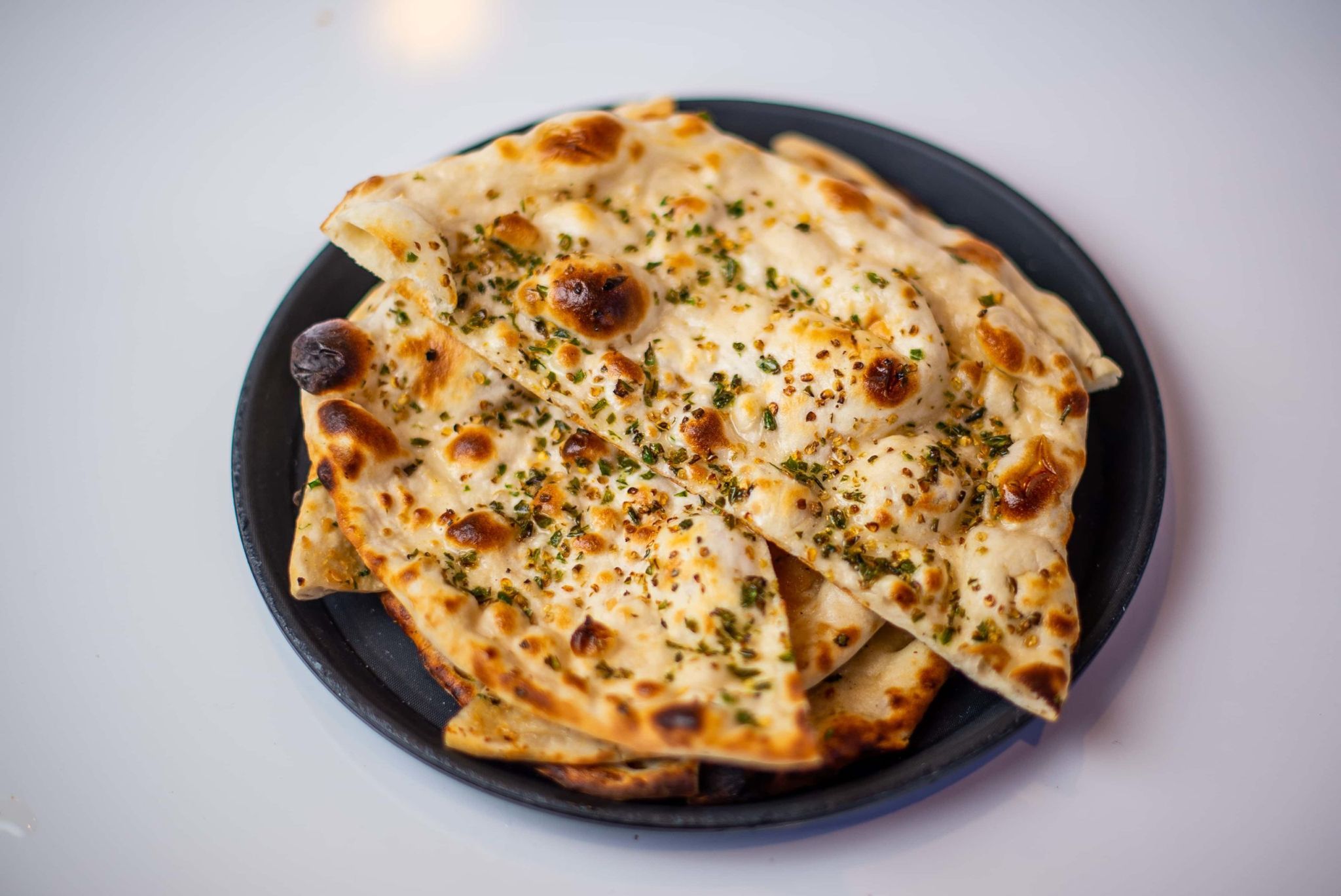 Several pieces of hot naan bread, sprinkled with green herbs and spices, on a black plate.