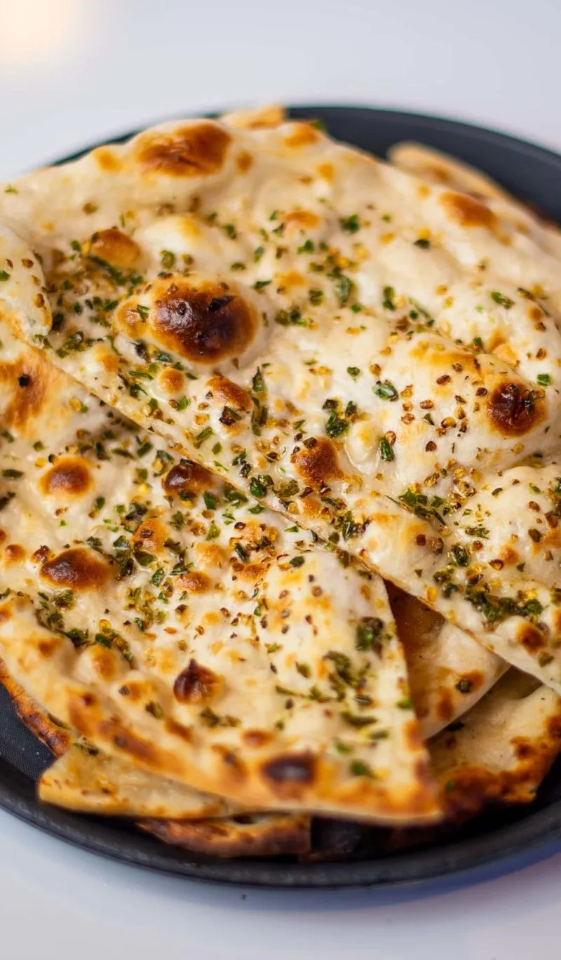 Several pieces of hot naan bread, sprinkled with green herbs and spices, on a black plate.
