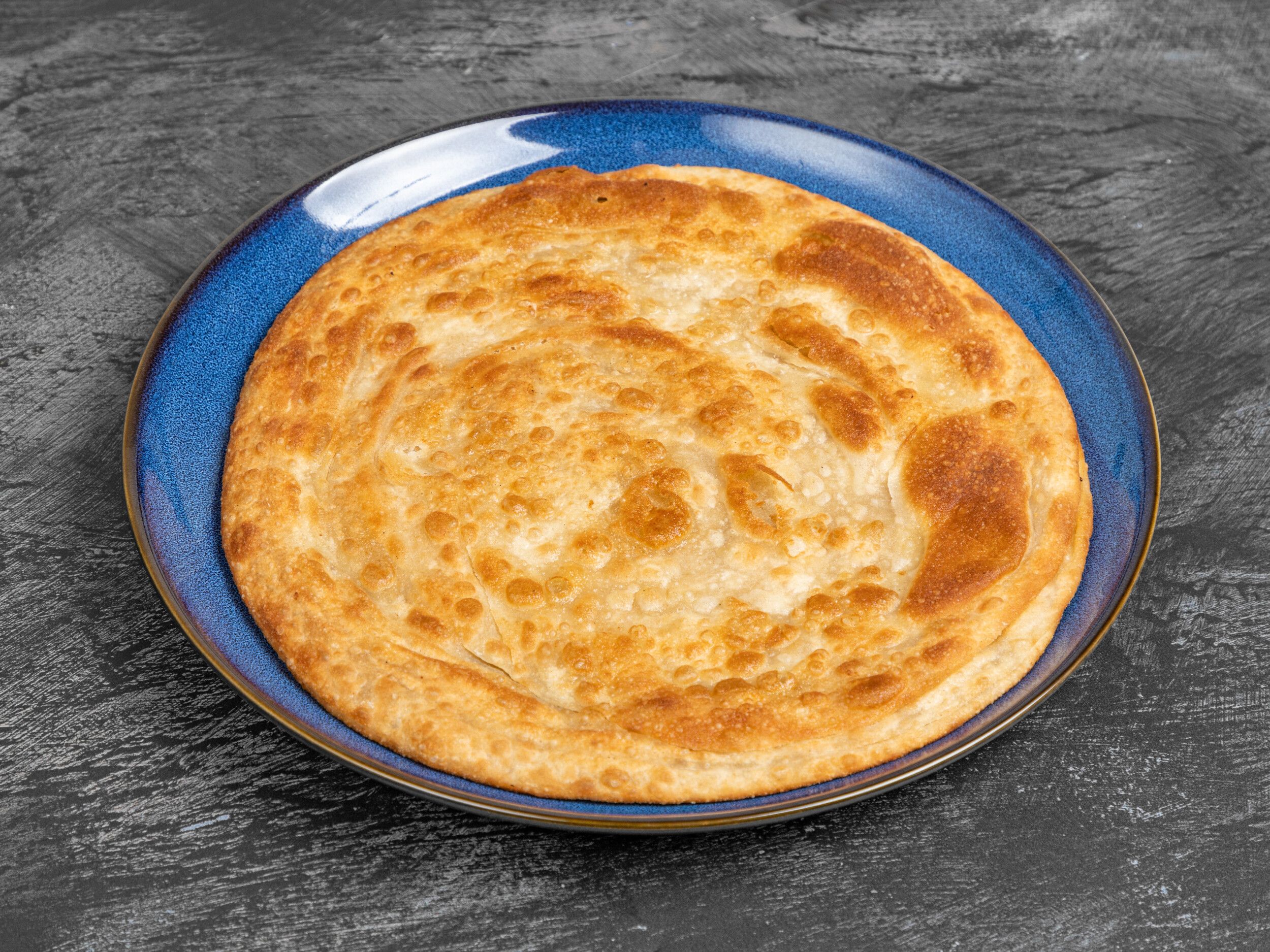 A golden, flaky paratha flatbread served on a glossy blue plate against a dark, textured background.