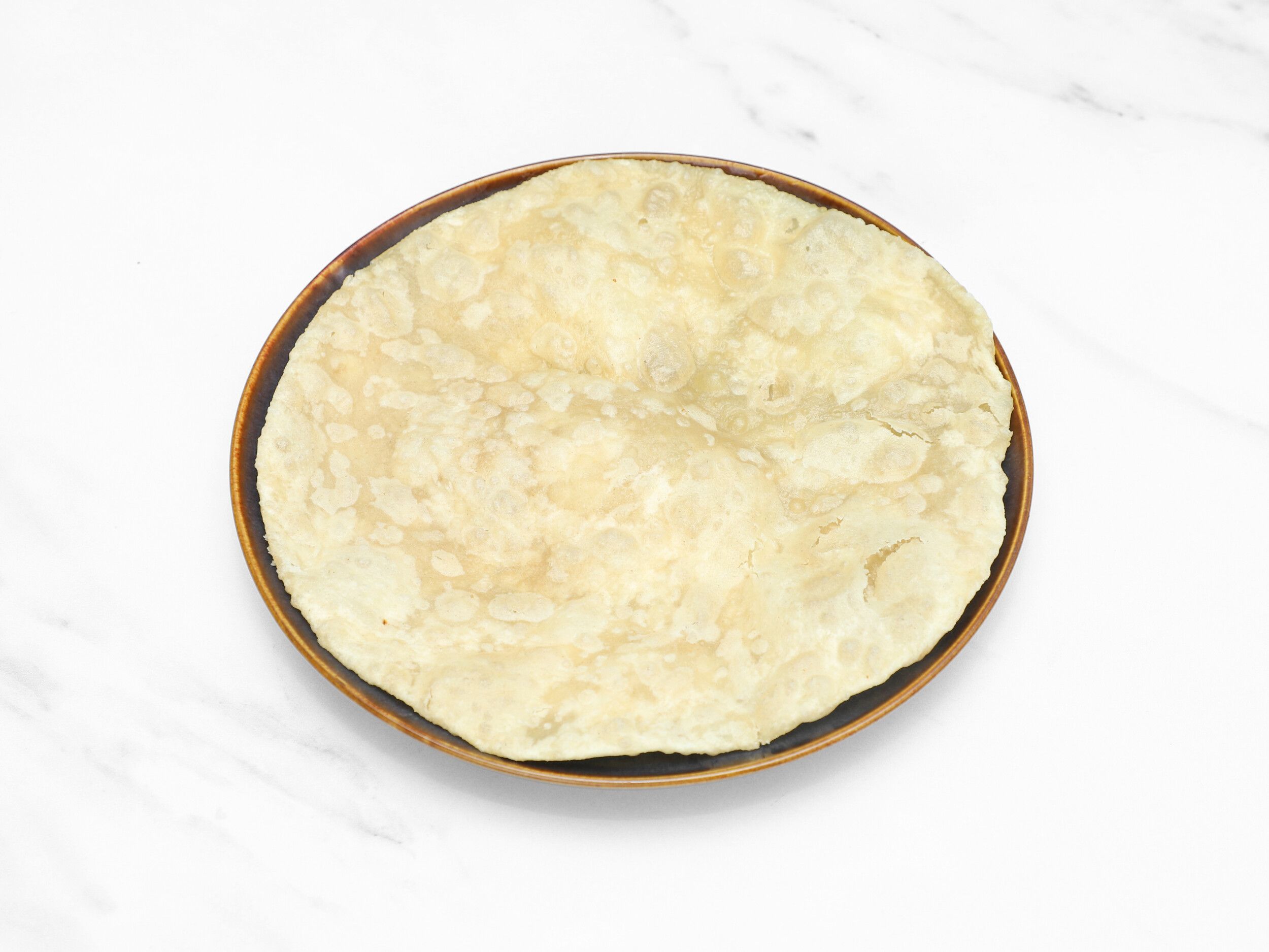 A round, light-colored Indian flatbread, possibly puri or paratha, resting on a brown plate.