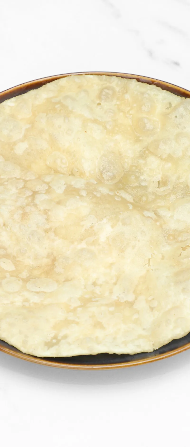A round, light-colored Indian flatbread, possibly puri or paratha, resting on a brown plate.