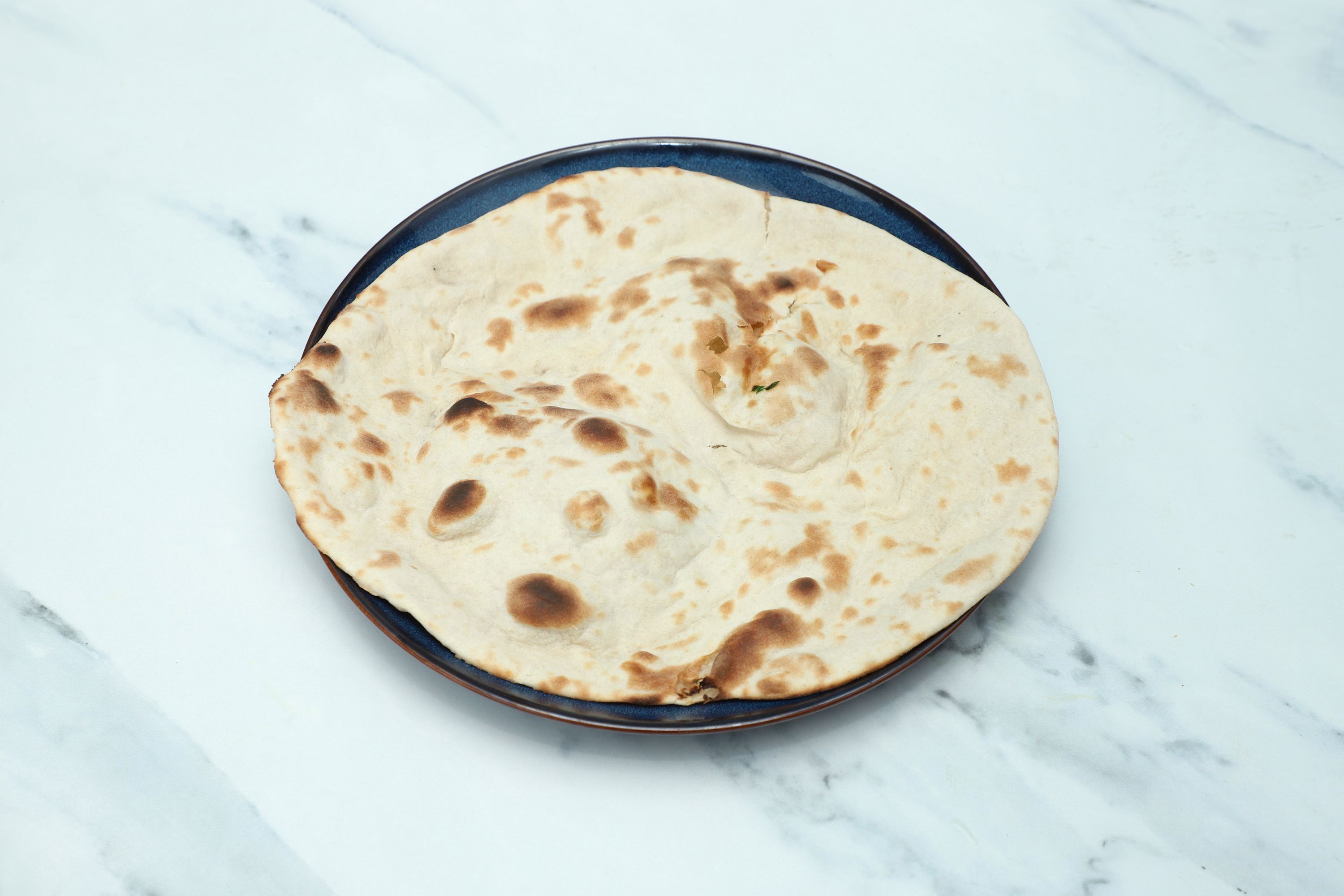 Round, golden-brown naan bread with slight charring, served on a dark blue plate on a marble surface.
