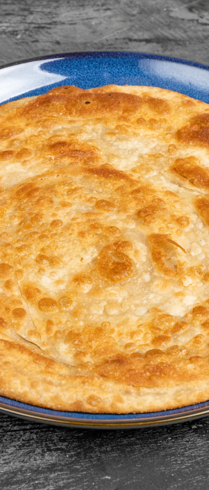 Round, golden-brown paratha bread with a flaky texture, served on a textured blue plate.