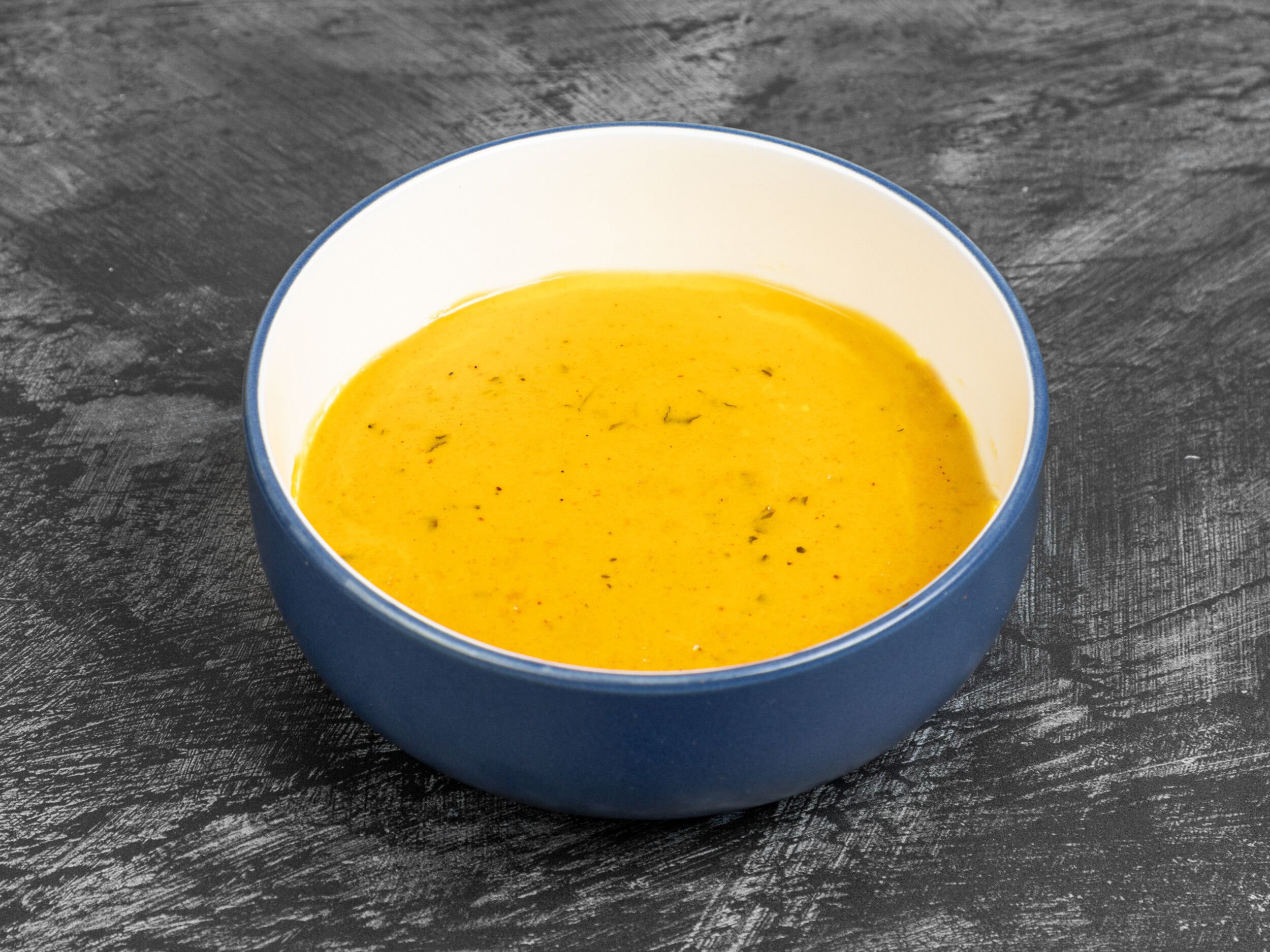 A round, blue-and-white bowl, filled with a smooth, orange-yellow soup with specks of herbs.