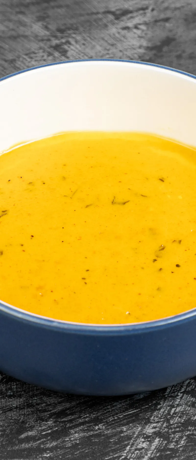 A round, blue-and-white bowl, filled with a smooth, orange-yellow soup with specks of herbs.