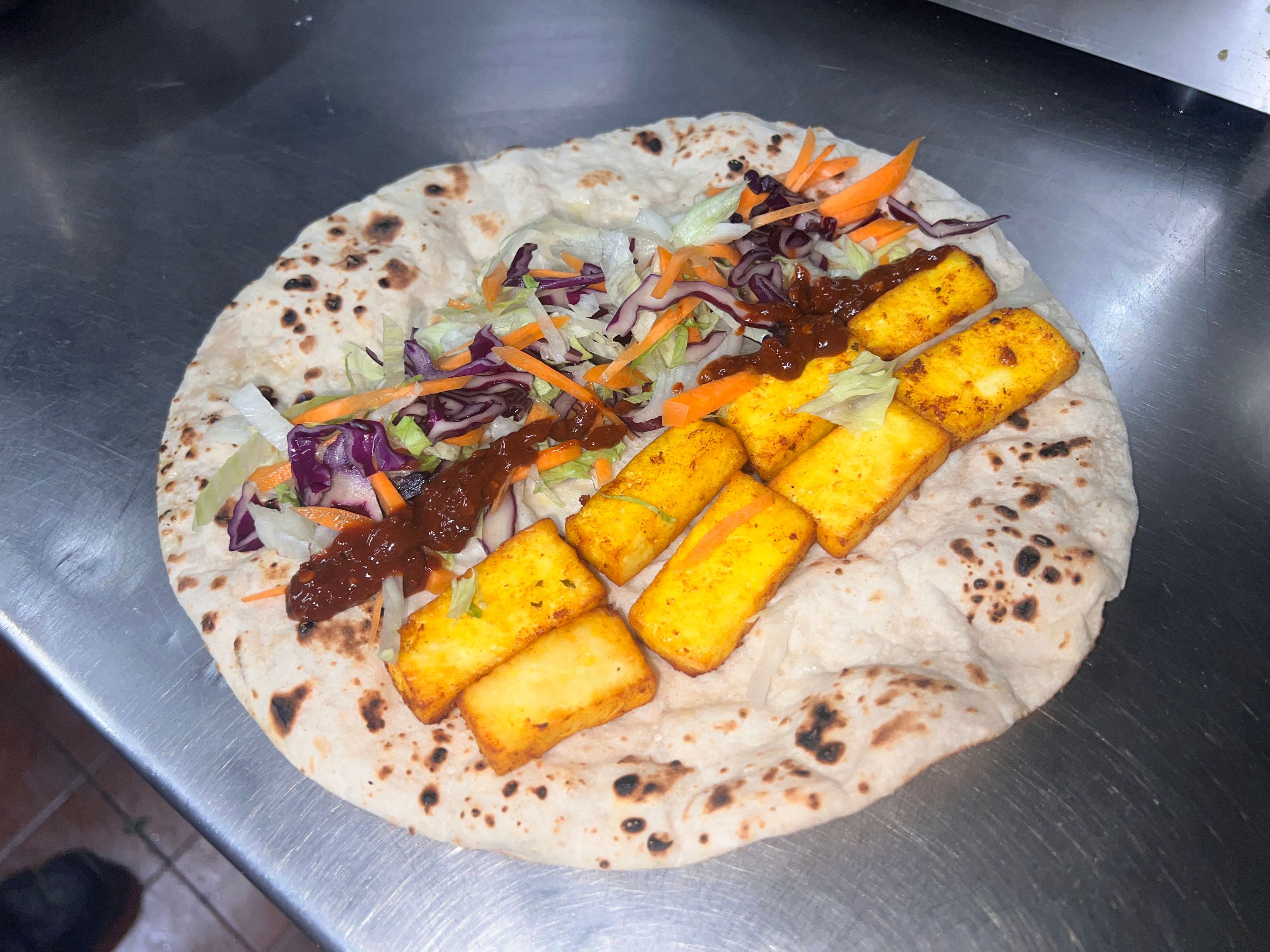 A flatbread wrap with golden fried paneer pieces, shredded lettuce, red cabbage, carrots, and a dark chutney.