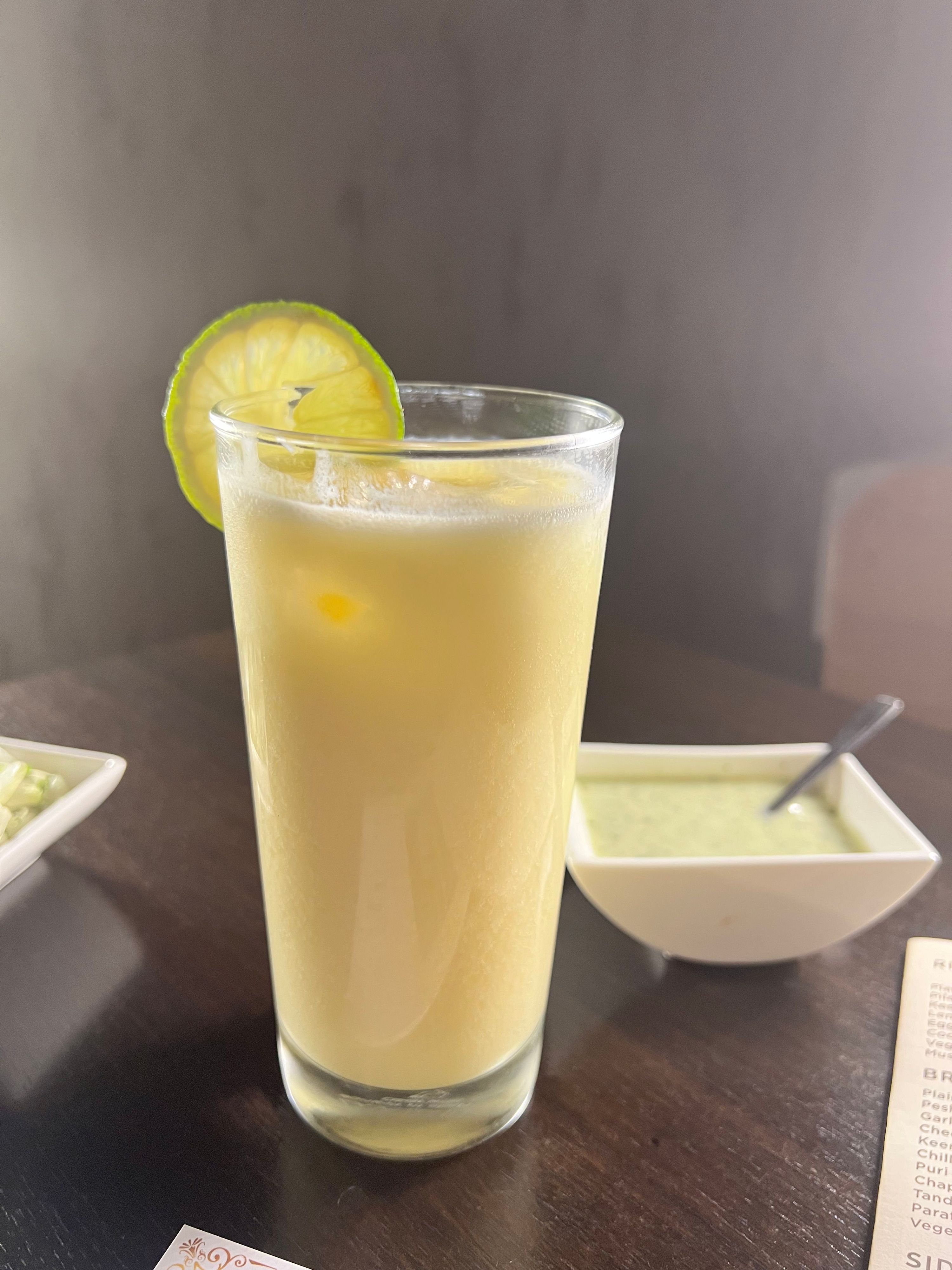 Mango lassi in a tall glass, garnished with a lime slice, next to a small bowl of green chutney.