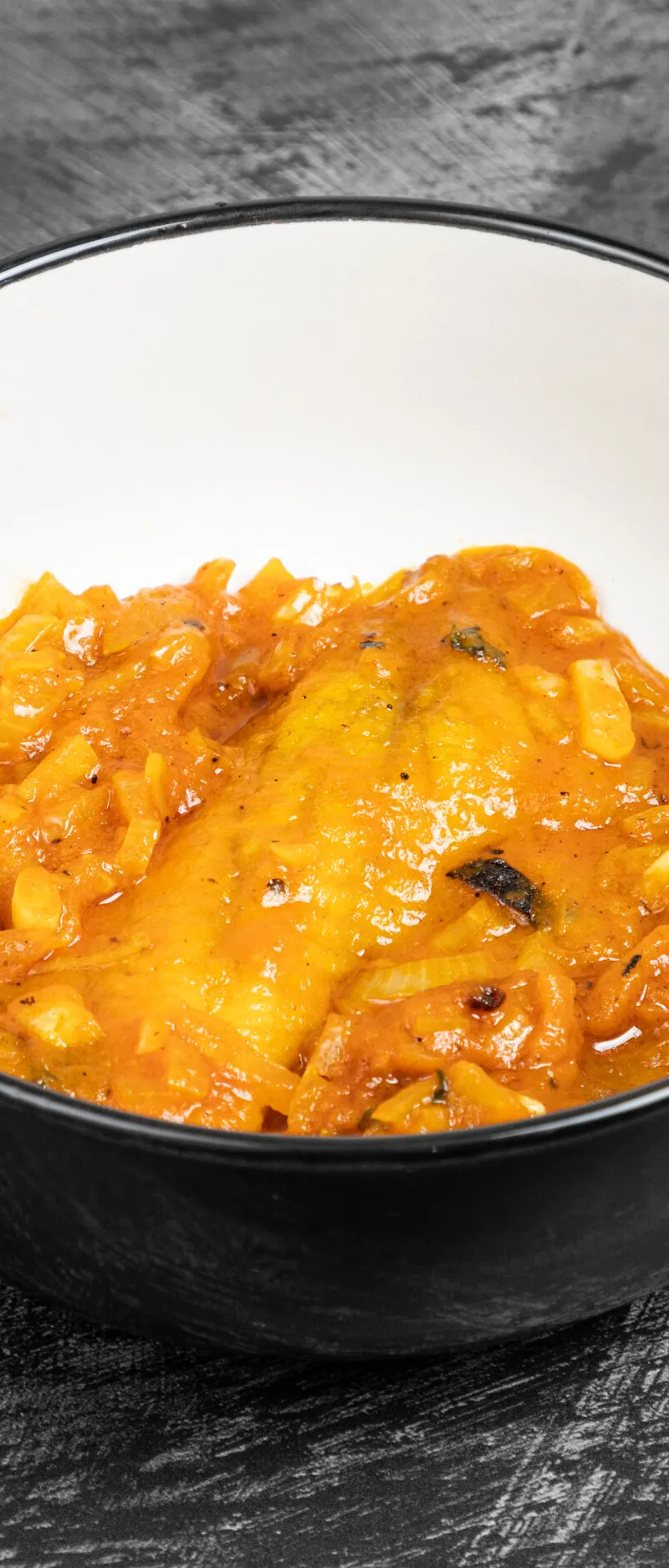 A close-up shot of a rich, orange-hued Indian curry with onions and spices in a white bowl with a black rim