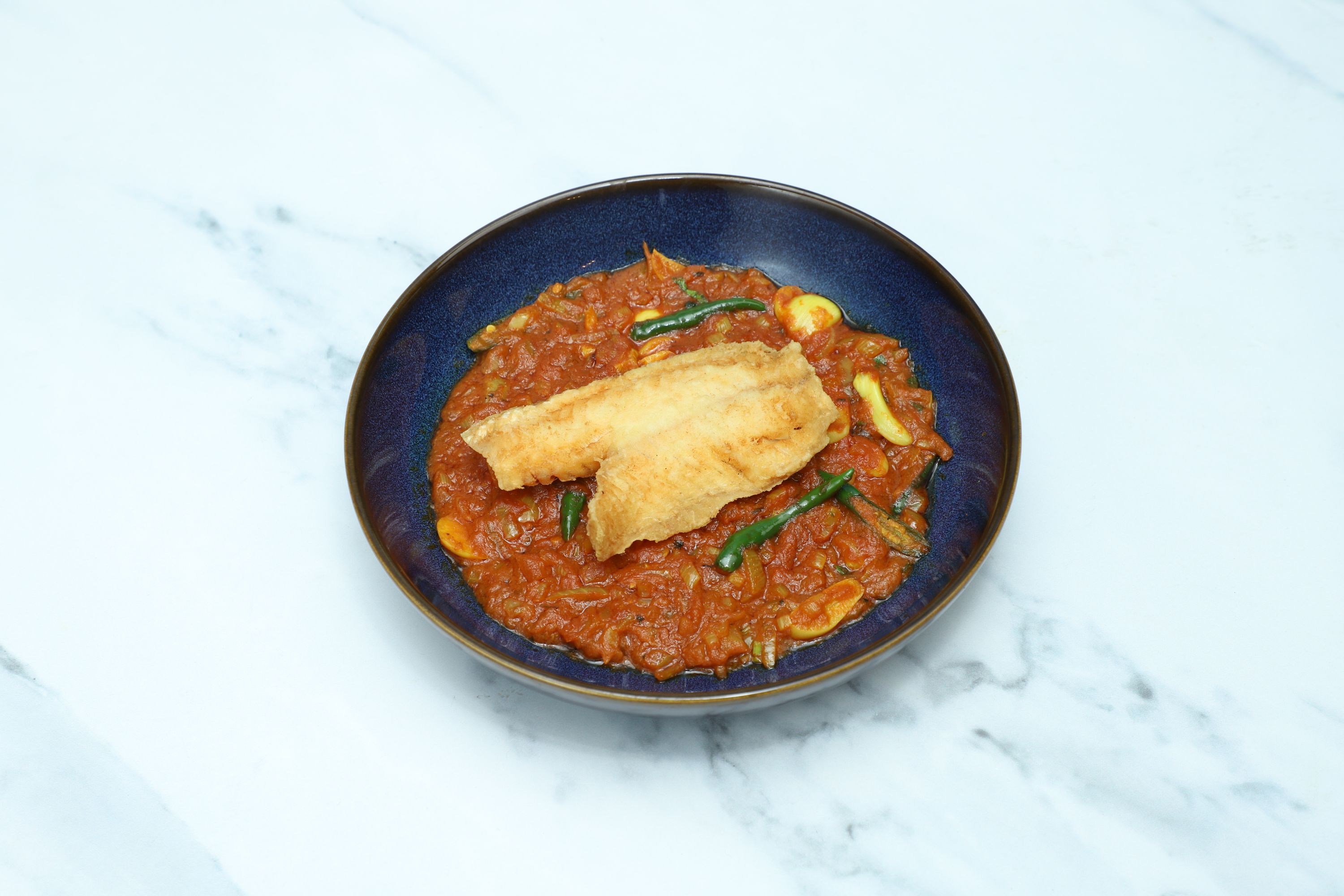 A crispy fried fish fillet rests atop a rich red tomato curry with green chilies in a dark blue bowl.
