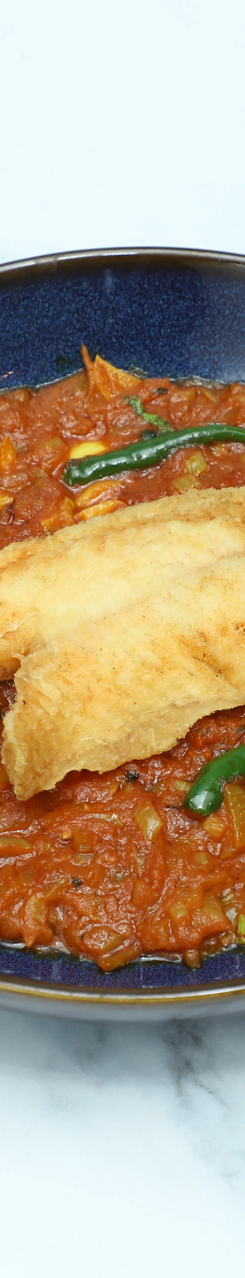 A crispy fried fish fillet rests atop a rich red tomato curry with green chilies in a dark blue bowl.