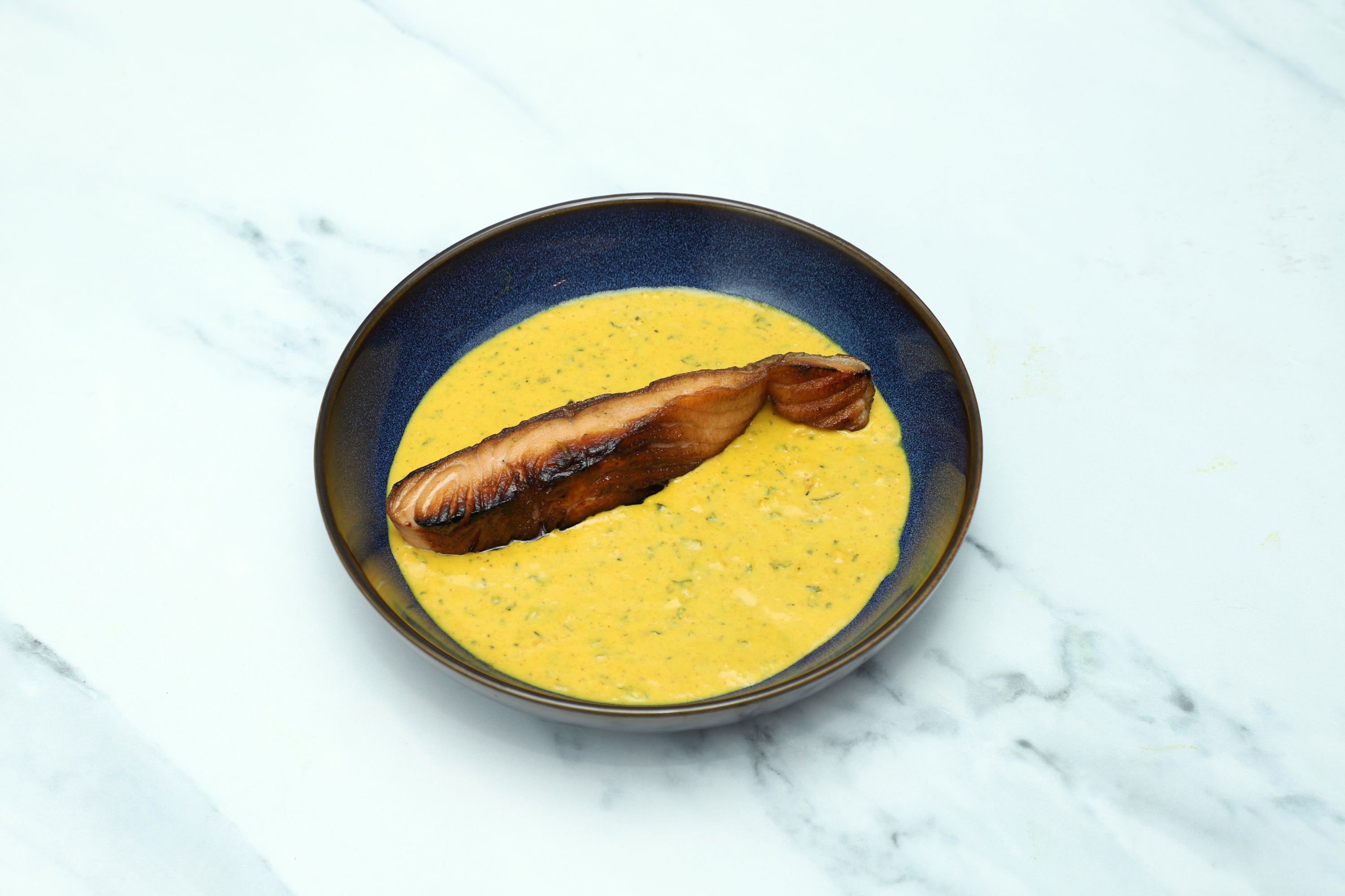 Grilled salmon fillet resting in a vibrant saffron-colored sauce with herbs in a dark-rimmed bowl.