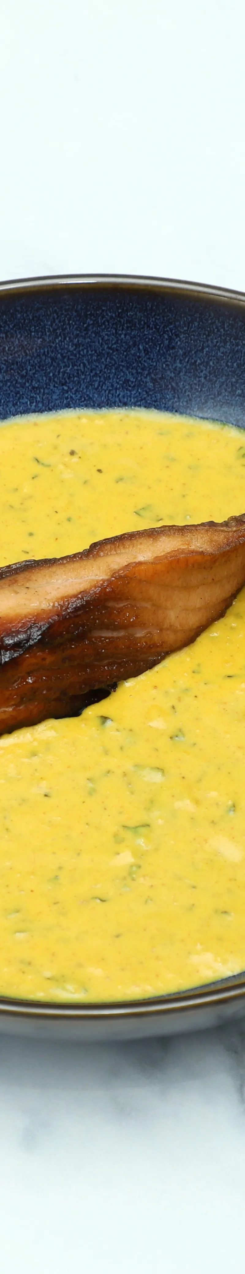 Grilled salmon fillet resting in a vibrant saffron-colored sauce with herbs in a dark-rimmed bowl.