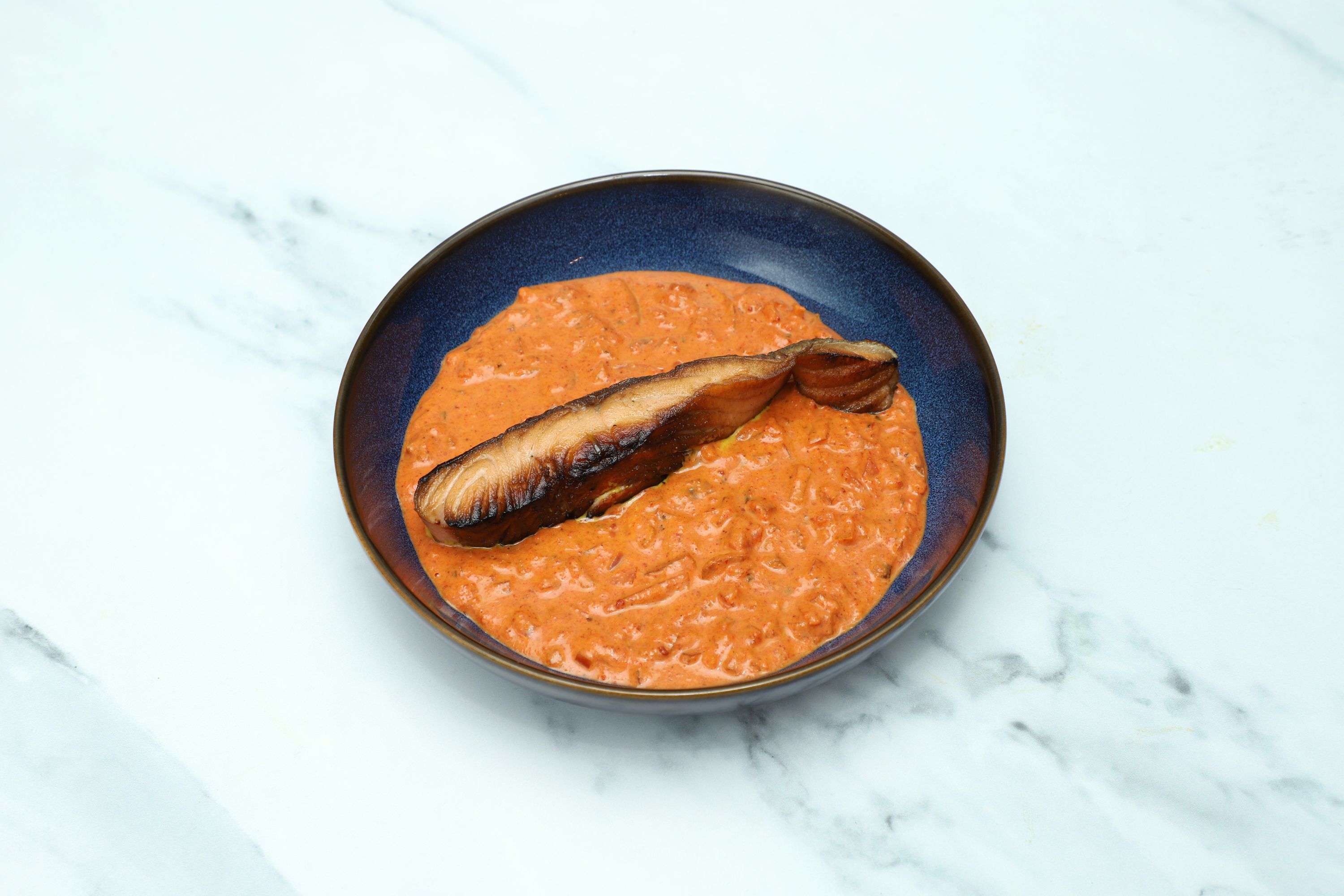 Grilled salmon fillet resting in a rich, creamy orange tomato-based curry in a blue bowl