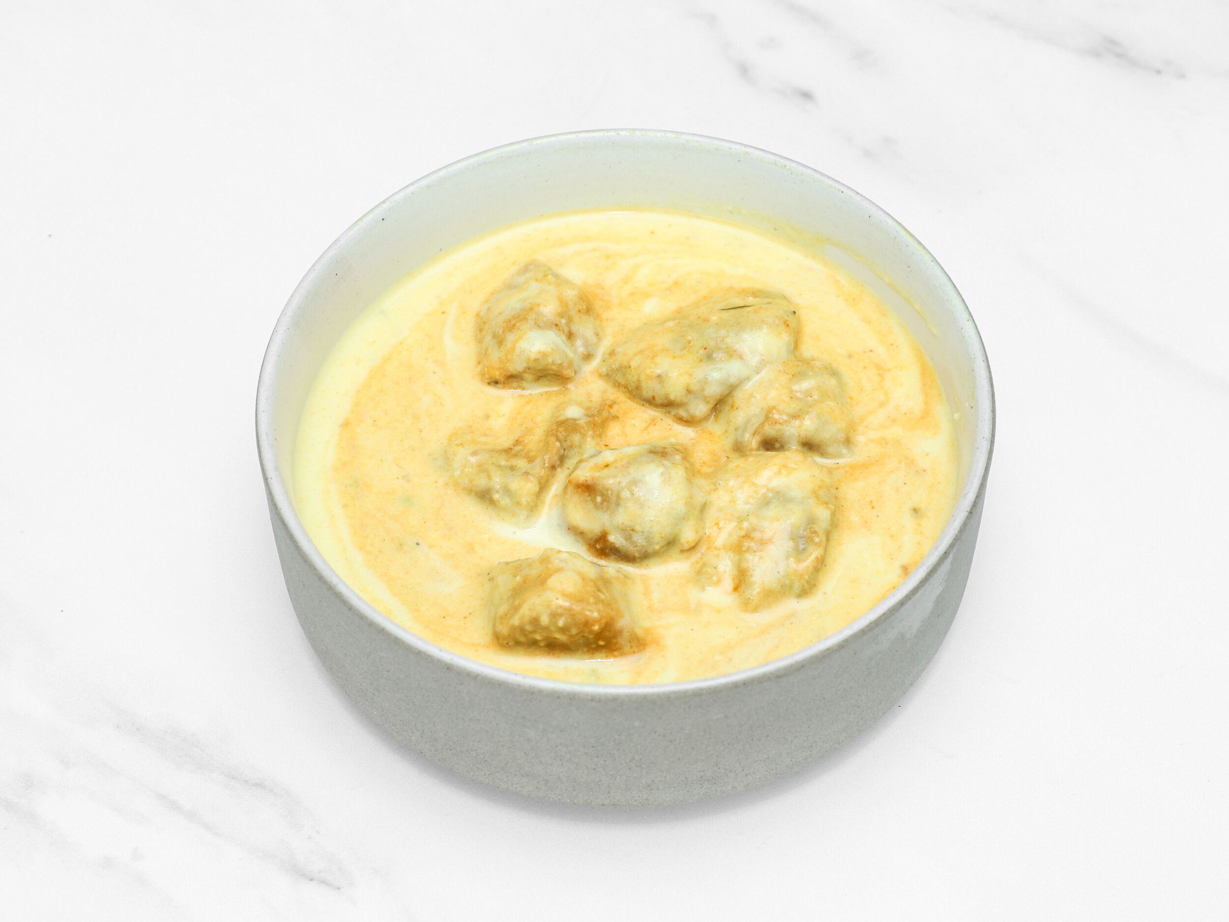 Malai Kofta dumplings submerged in a rich, creamy yellow sauce, presented in a light gray bowl.