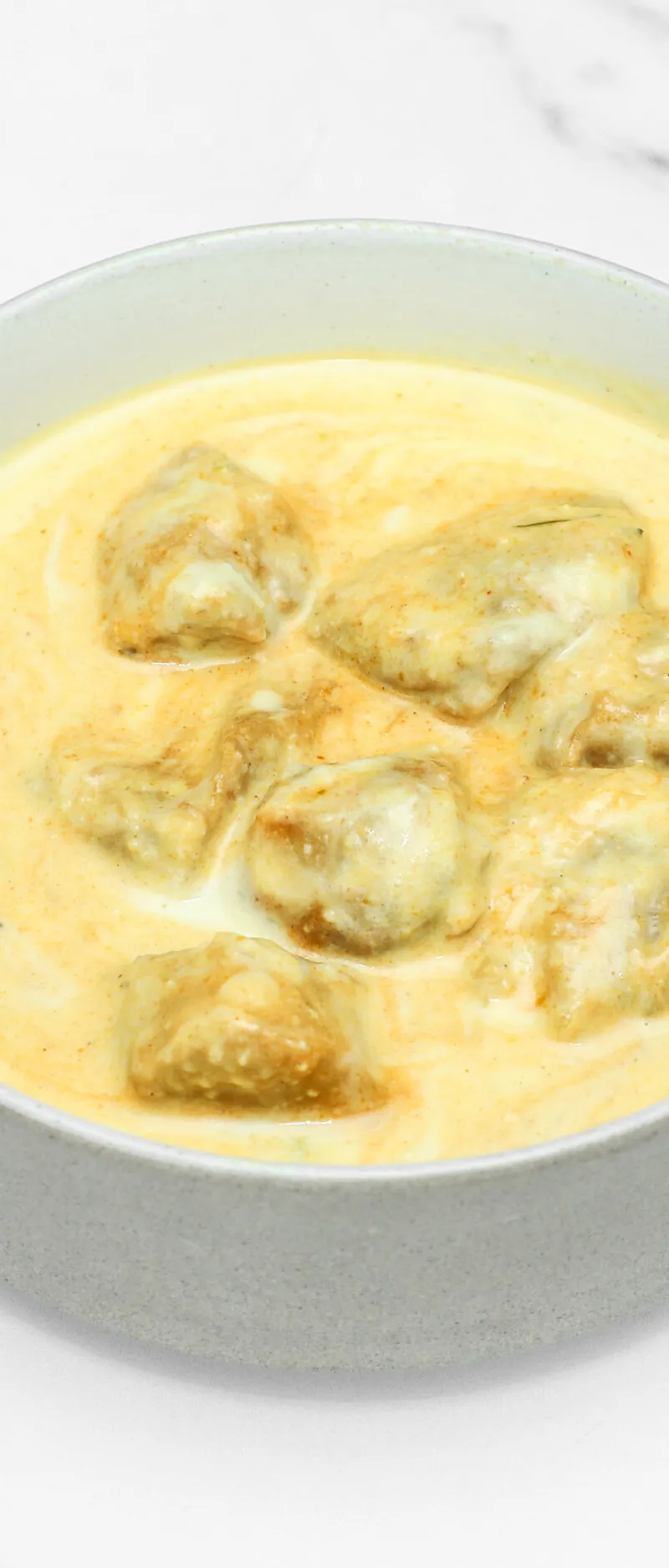 Malai Kofta dumplings submerged in a rich, creamy yellow sauce, presented in a light gray bowl.